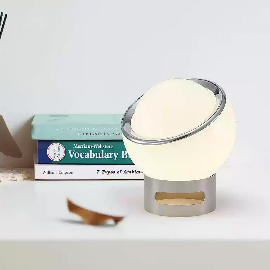 White Clérachic table lamp tilted slightly forward on a white shelf, with a stack of books and a dry leaf beside it.