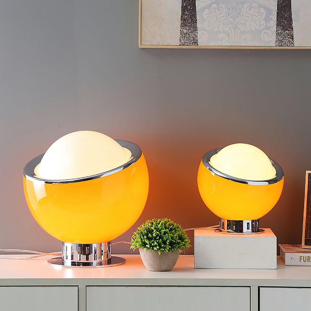 Two orange Clérachic table lamps, one large and one small, lit on a white sideboard beside a small potted plant.