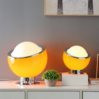 Two orange Clérachic table lamps, one large and one small, lit on a white sideboard beside a small potted plant.
