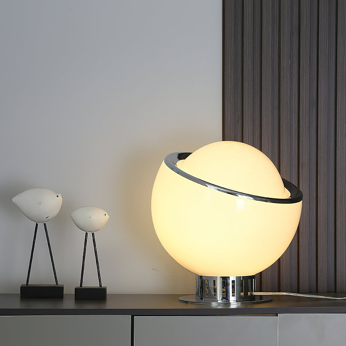 Large white glass globe Clérachic table lamp on a dark sideboard, with two white bird figures and a slatted wall behind.