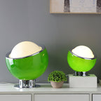 Two green Clérachic table lamps, a large one and a small one, lit on a white sideboard beside a potted plant against a grey wall.