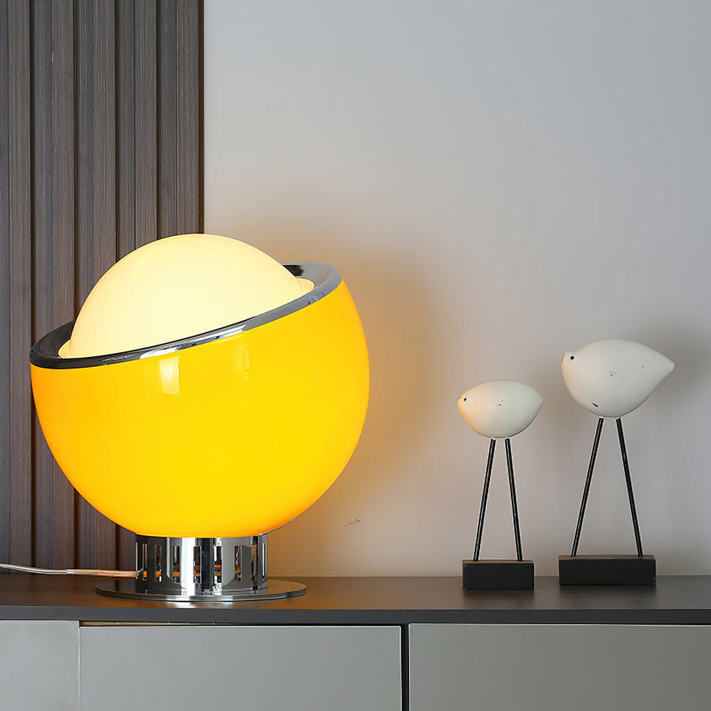 Yellow-toned glass Clérachic table lamp lit on a dark shelf beside two white bird figures, with a slatted panel behind.