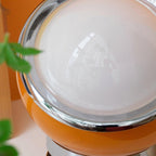 Close view of the white glass dome, chrome rim and orange outer shell of this Clérachic table lamp, with a plant leaf in the corner.