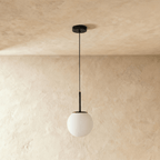 Clérachic Cells pendant light, Straight profile in Black, its frosted globe glowing softly against a warm plaster wall.