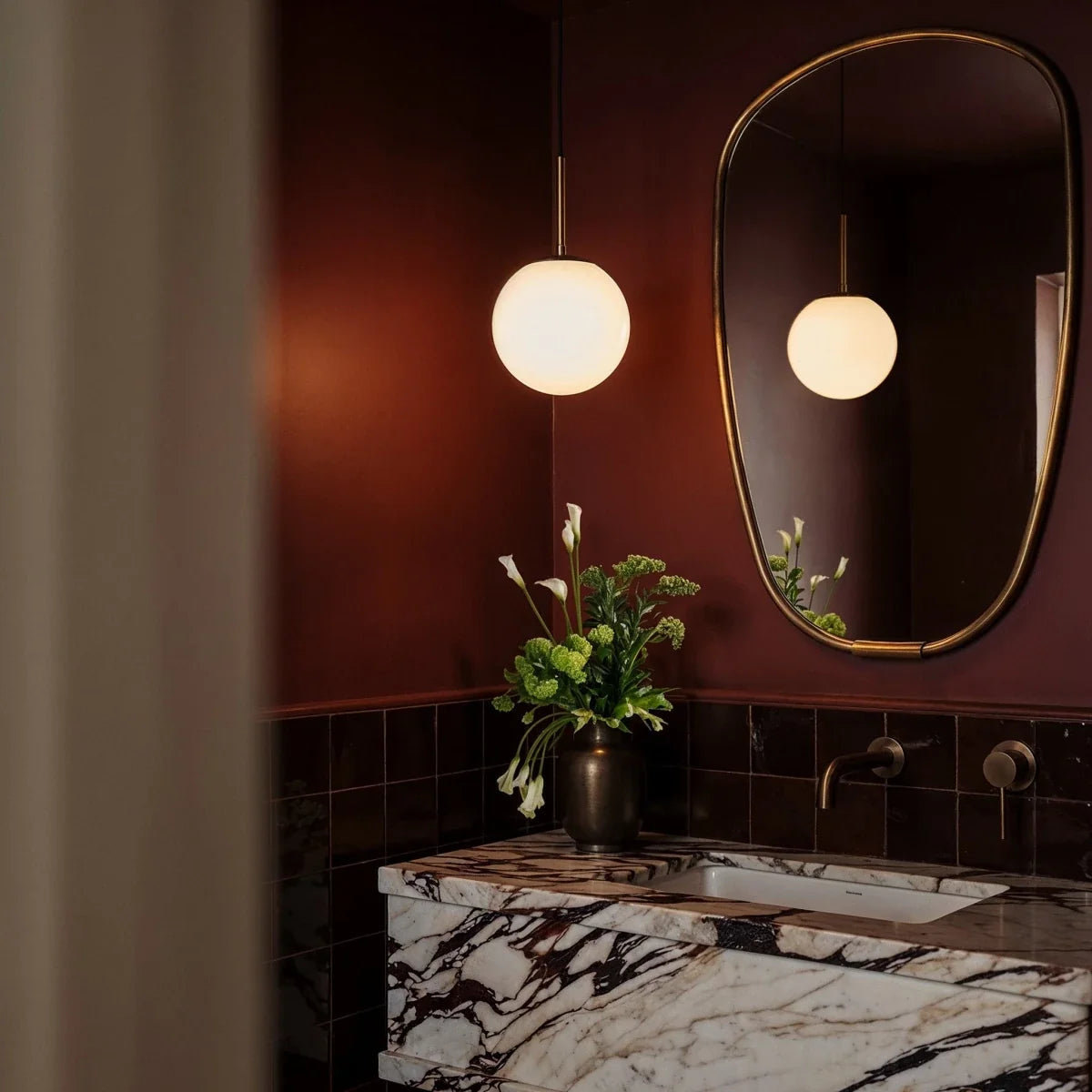 Clérachic Cells pendant light in Straight, Brass, glowing beside a marble vanity in a deep red-tiled powder room.