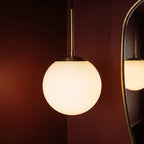 Close-up of the Clérachic Cells pendant light in Straight, Brass, its frosted globe lit warm against a red wall.