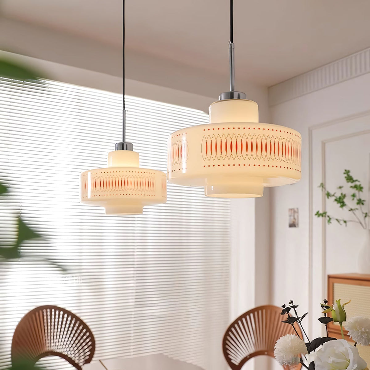 Two Clérachic pendants of different sizes hang by a sheer-blinded window, with fan-back chairs and white flowers below.