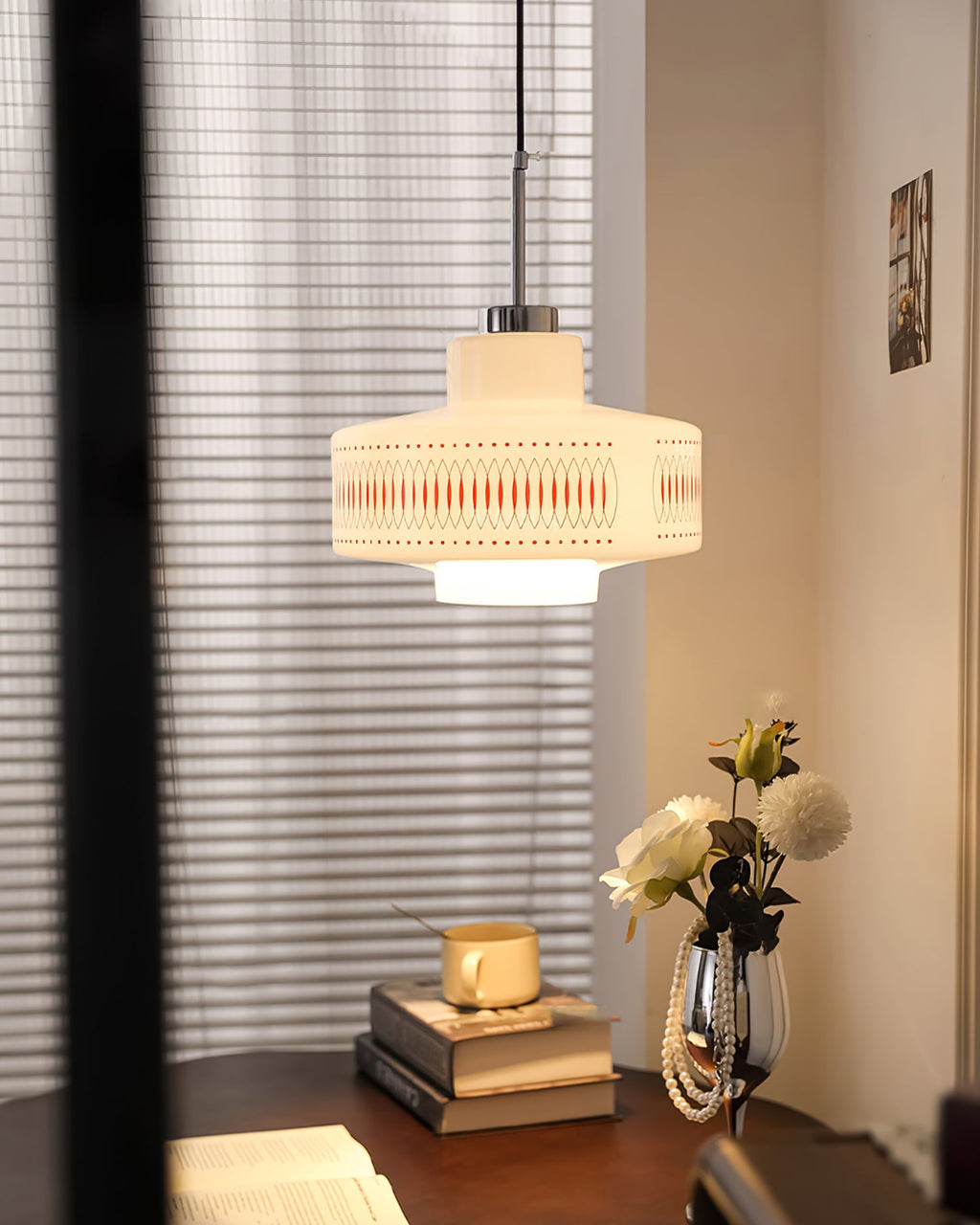 A Clérachic pendant glows in a corner beside a sheer blind, with a dark side table, books and white flowers below.