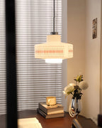 A Clérachic pendant glows in a corner beside a sheer blind, with a dark side table, books and white flowers below.
