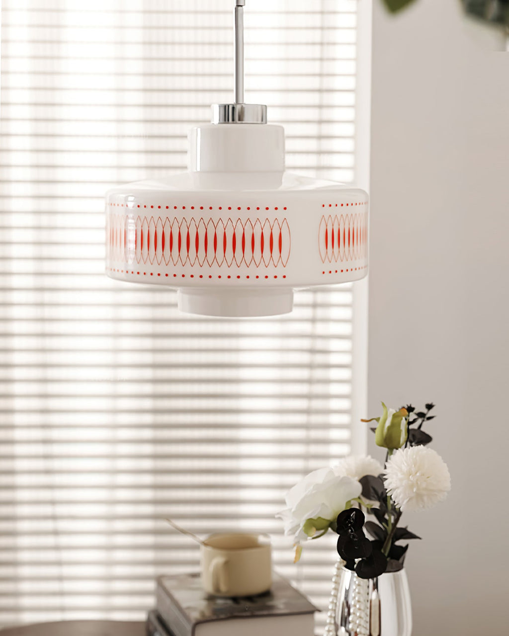 Clérachic pendant with a white glass shade and orange diamond print, hung in front of a blind with white peonies beneath.