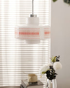 Clérachic pendant with a white glass shade and orange diamond print, hung in front of a blind with white peonies beneath.