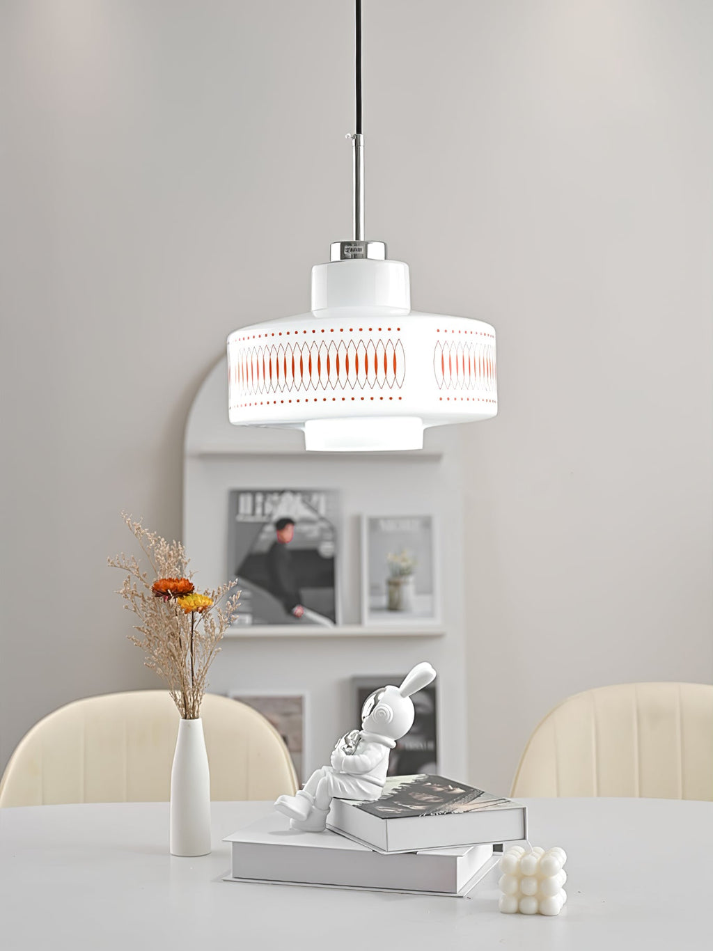 A Clérachic pendant with a white glass drum and orange print hangs above a white table with a vase and a rabbit figure.