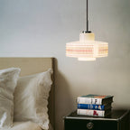 A Clérachic pendant hangs above a bedside table with stacked books, beside a bed with white broderie anglaise pillows.
