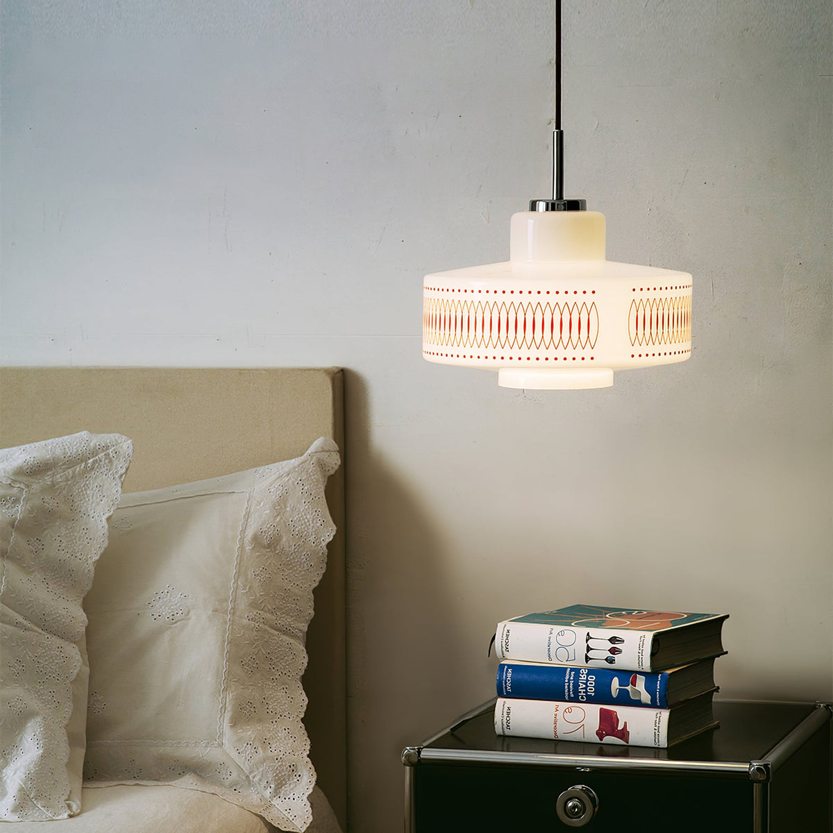 A Clérachic pendant hangs above a bedside table with stacked books, beside a bed with white broderie anglaise pillows.