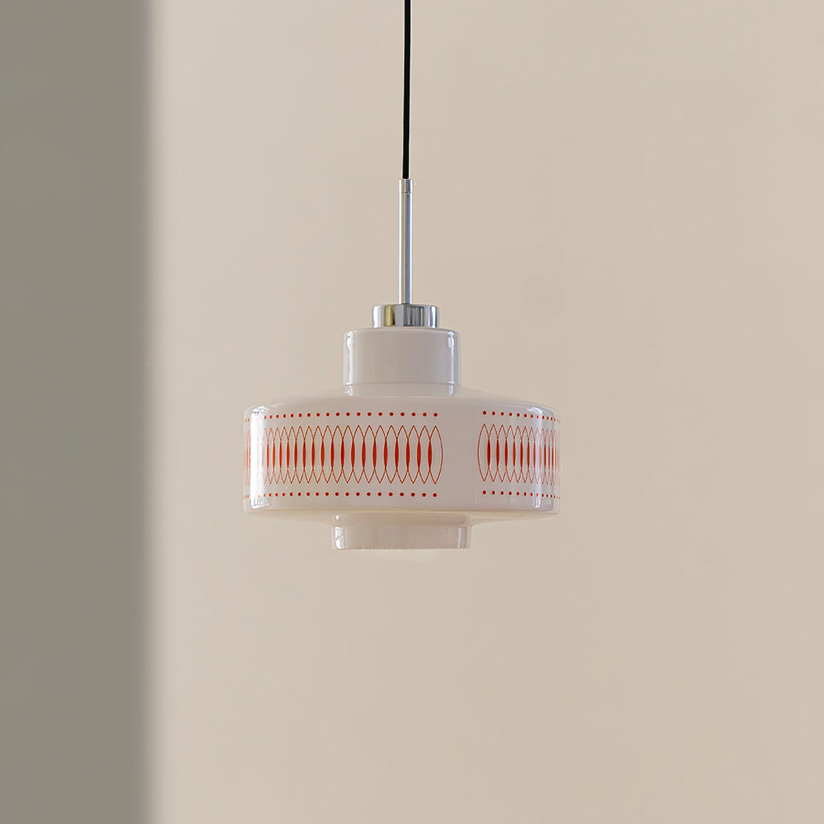 Clérachic pendant with a white glass shade, orange lattice print and a chrome stem, hung against a tan and grey wall.