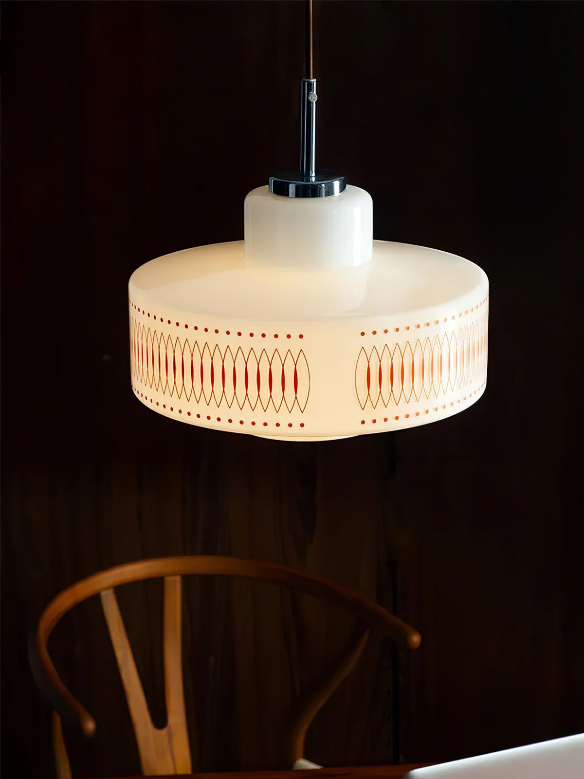 A Clérachic pendant glows against a dark wood wall, with the back of a wooden wishbone chair in the corner.