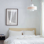 A Clérachic pendant hangs high in a white bedroom, with a framed print, a bed with white linen and a stack of books.