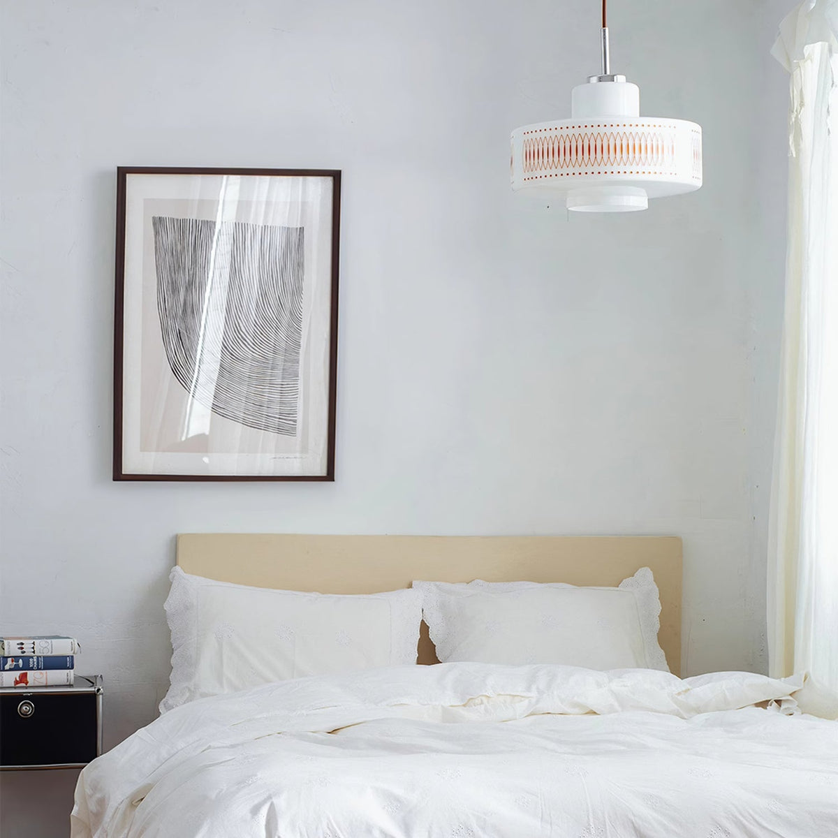 A Clérachic pendant hangs high in a white bedroom, with a framed print, a bed with white linen and a stack of books.