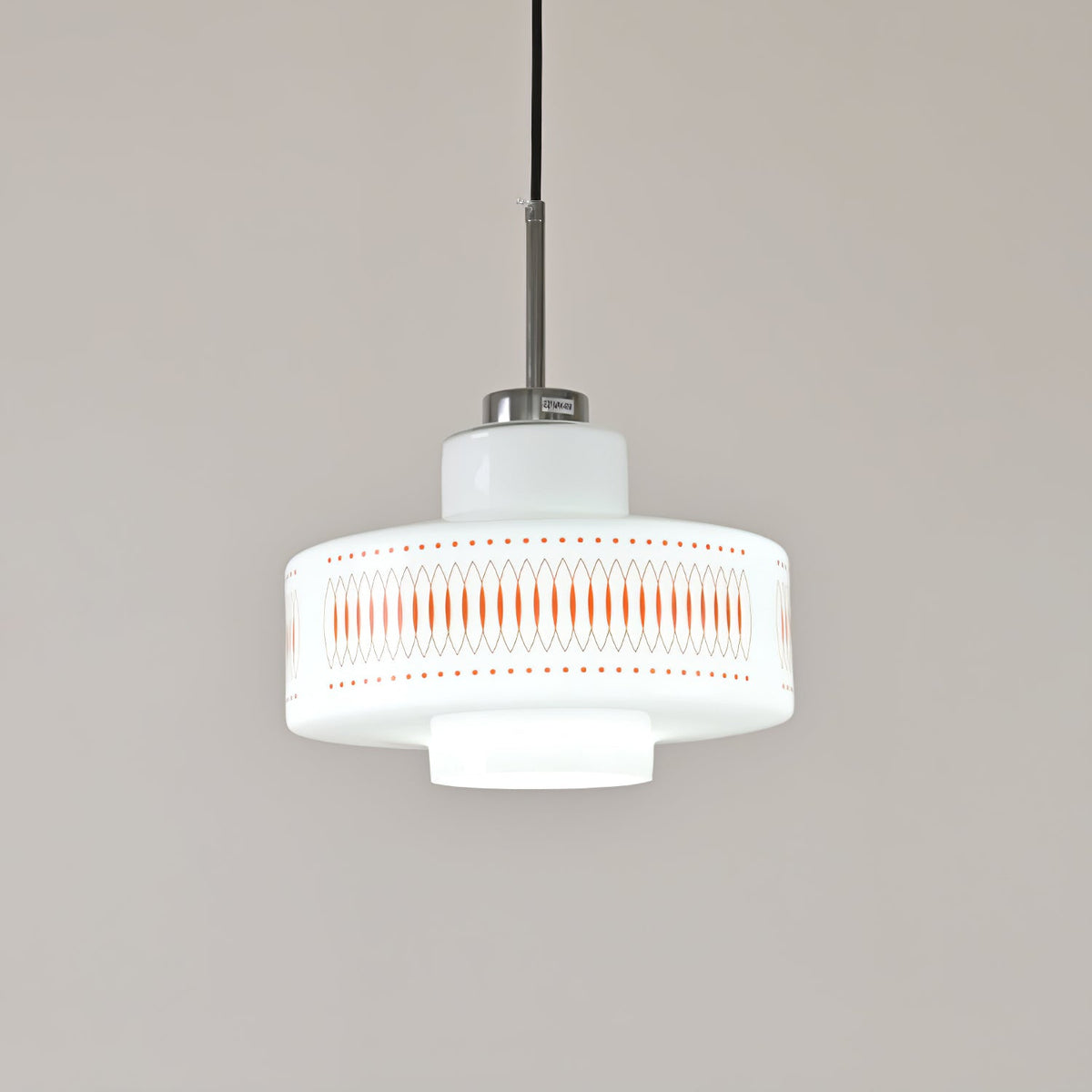 A Clérachic pendant in white opal glass with orange diamond print, shown in cool white light against a grey wall.