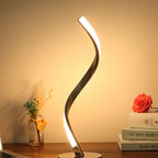 Clérachic spiral LED table lamp glowing warm on a wooden desk beside a small vase of dried flowers and a stack of books.