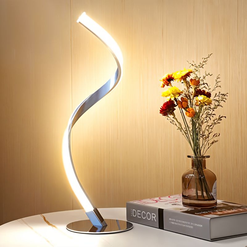 Clérachic chrome spiral LED table lamp lit warm on a white marble-top table beside a vase of flowers and a book.