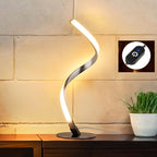 Clérachic spiral LED table lamp lit warm against a pale brick wall, with an inset of the inline touch switch on its cable.