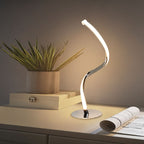 Clérachic polished chrome spiral LED table lamp glowing warm on a white sideboard beside a potted plant and an open magazine.