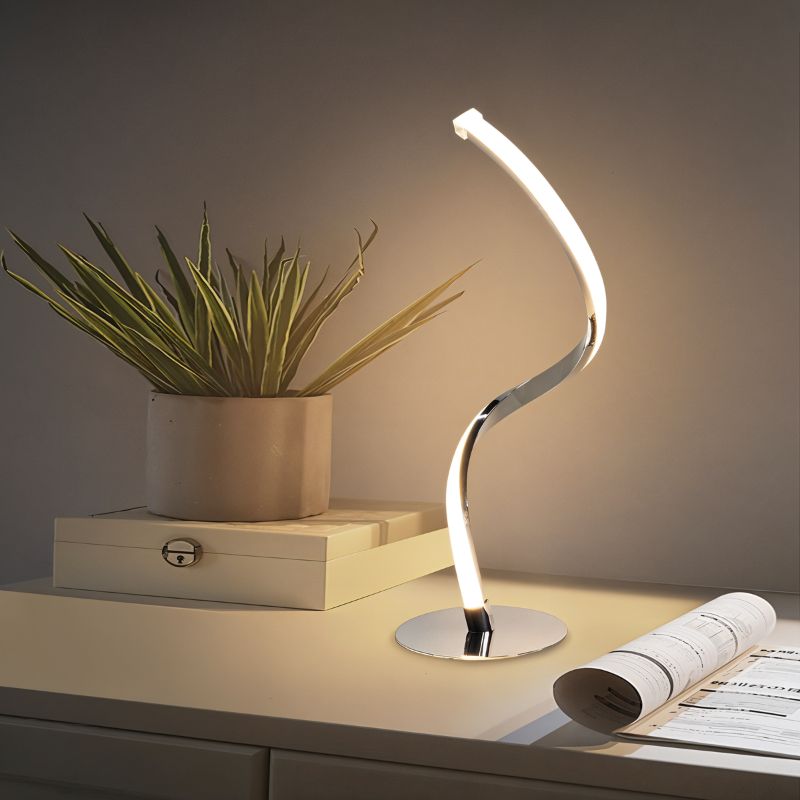 Clérachic polished chrome spiral LED table lamp glowing warm on a white sideboard beside a potted plant and an open magazine.