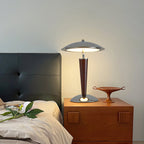 This Clérachic table lamp glows warmly on a wooden bedside cabinet, beside a bed with white linen and a dark leather headboard.