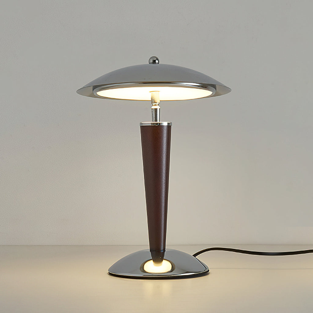 Front view of a lit Clérachic table lamp on a pale surface, its chrome dome and walnut stem standing against a soft grey wall.