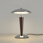 Front view of a lit Clérachic table lamp on a pale surface, its chrome dome and walnut stem standing against a soft grey wall.