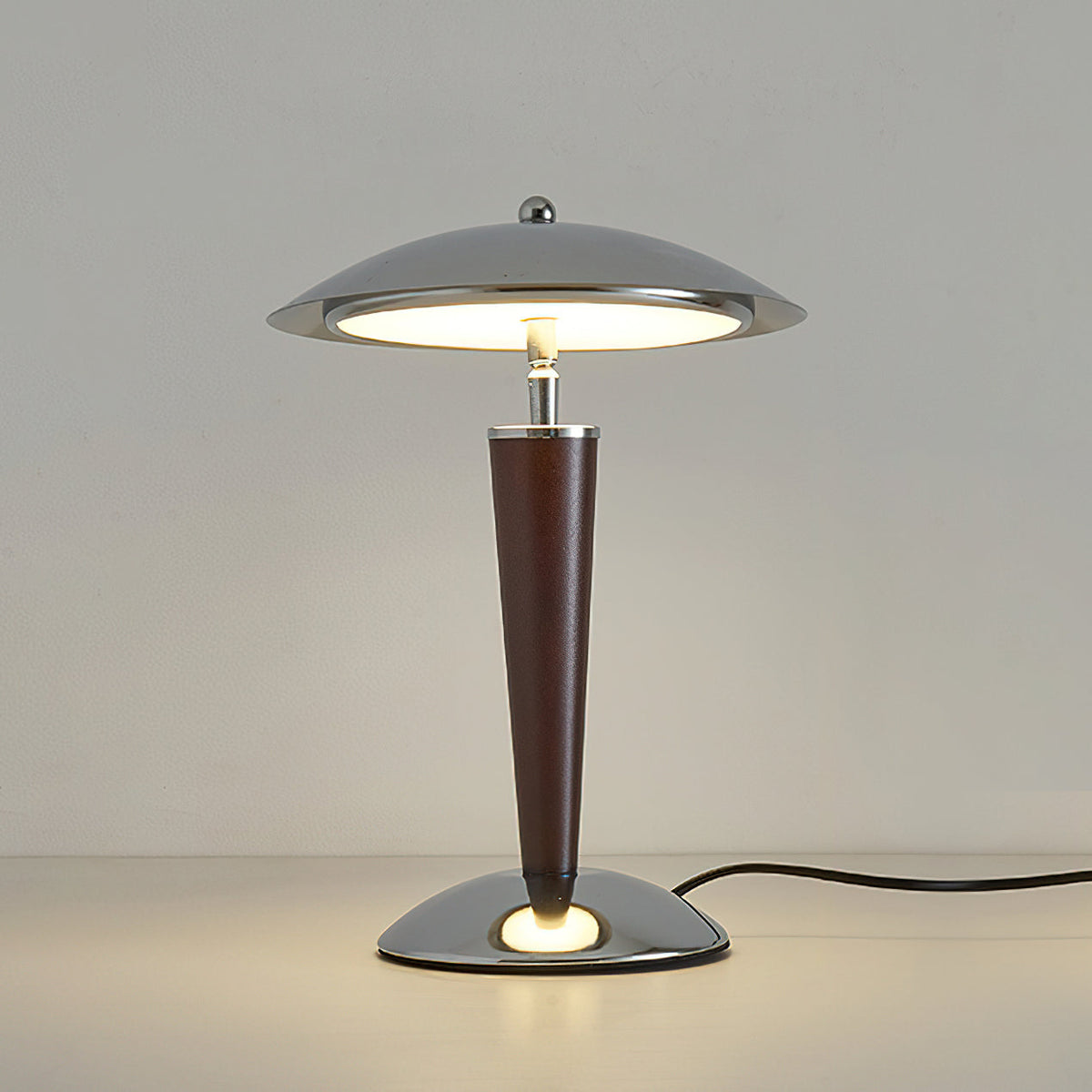 Front view of a lit Clérachic table lamp on a pale surface, its chrome dome and walnut stem standing against a soft grey wall.