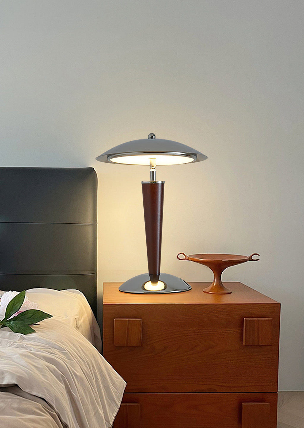 Small Clérachic table lamp lit on a wooden bedside cabinet, with the corner of a bed and a green leaf in view.