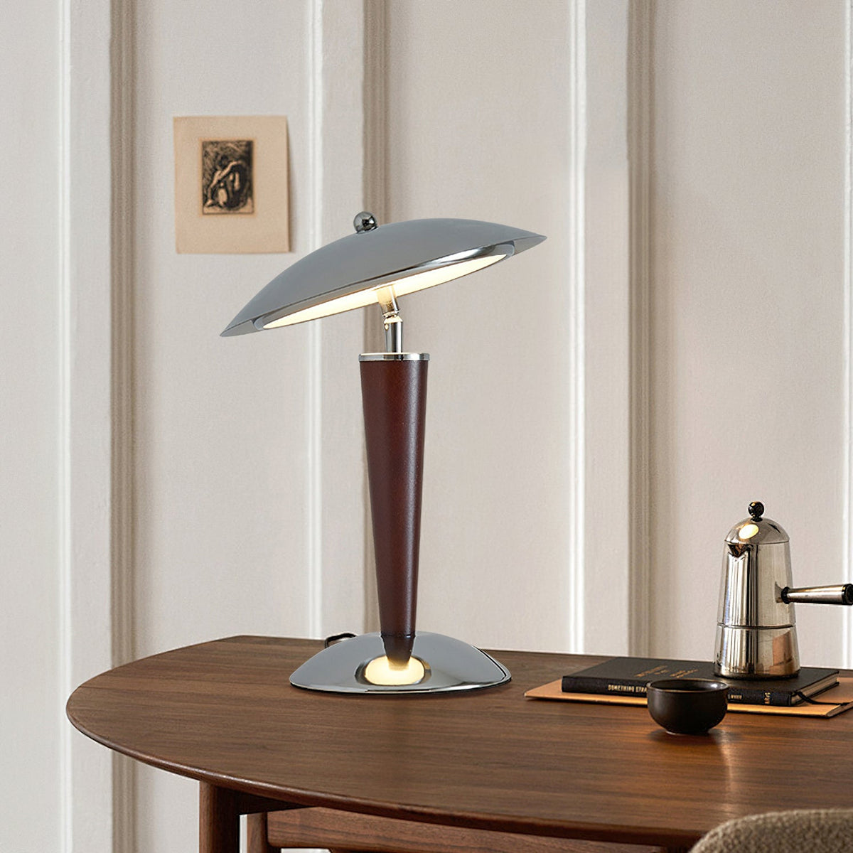 Angled view of a Clérachic chrome dome lamp on a curved walnut table, with a stovetop coffee pot and a small dark bowl beside it.