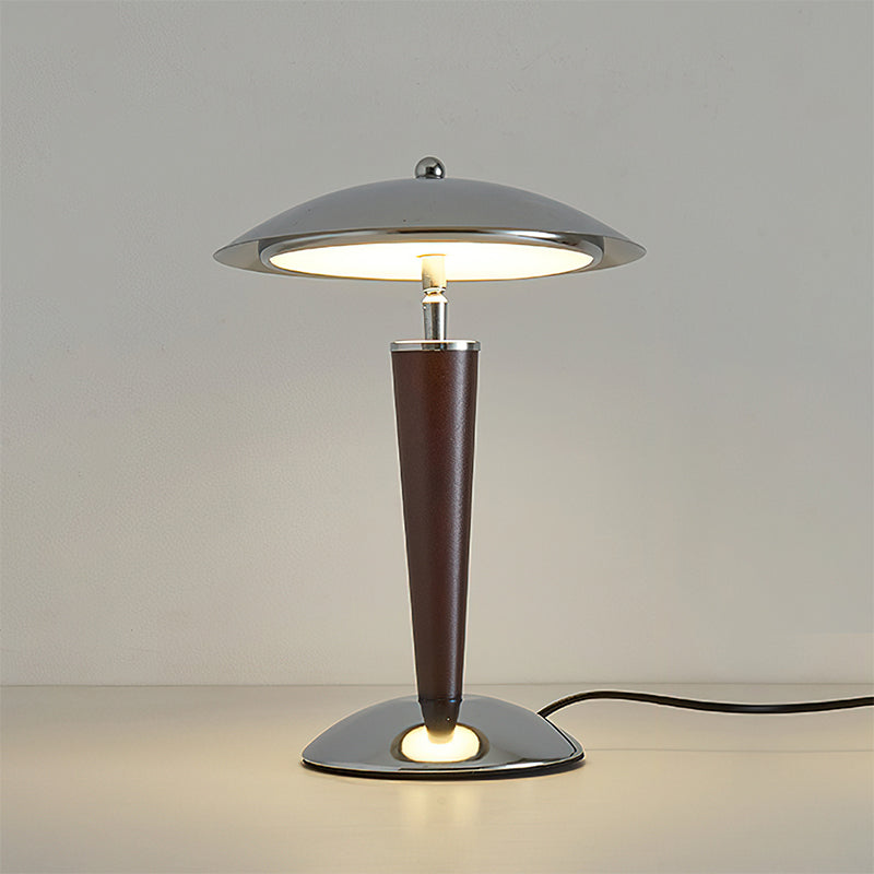 Straight-on view of a Clérachic table lamp with its black cord trailing to the right, lit against a plain grey wall.