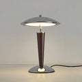 Straight-on view of a Clérachic table lamp with its black cord trailing to the right, lit against a plain grey wall.