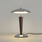 Straight-on view of a Clérachic table lamp with its black cord trailing to the right, lit against a plain grey wall.