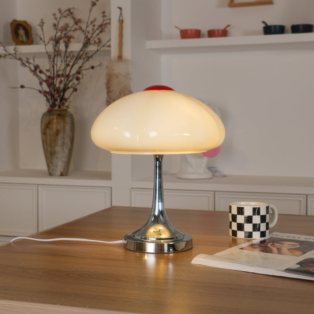 Warm light glows through the white scalloped shade of this Clérachic lamp, on a wooden desk with a checked mug and a magazine.