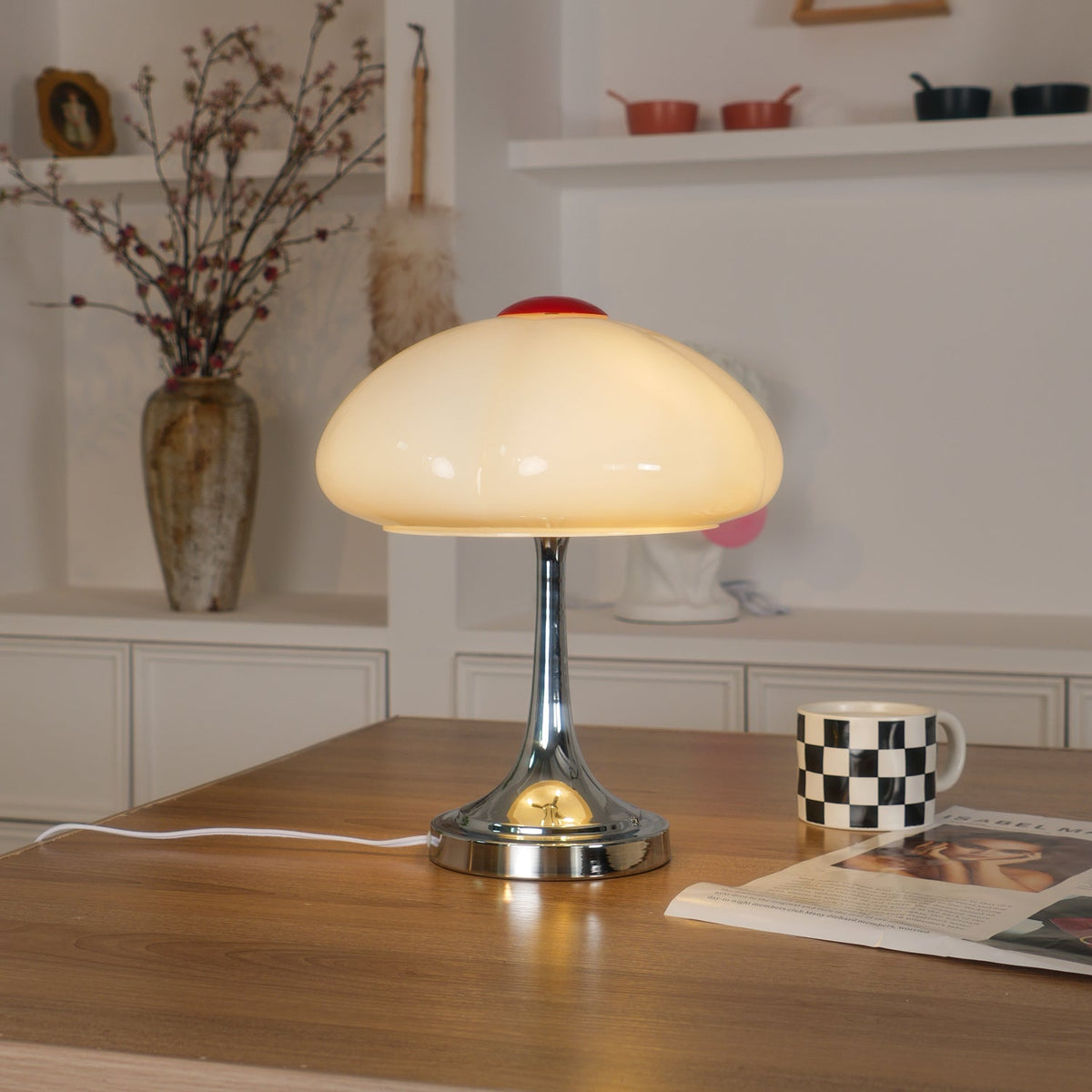 Warm light glows through the white scalloped shade of this Clérachic lamp, on a wooden desk with a checked mug and a magazine.