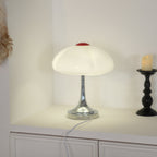 A Clérachic table lamp lit on a white shelf in a corner, its scalloped shade glowing above a chrome trumpet-shaped stem.