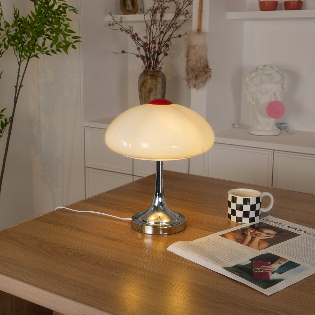 Lit Clérachic lamp on a wooden desk, a checked mug and open magazine beside it and a shelf of decor behind.