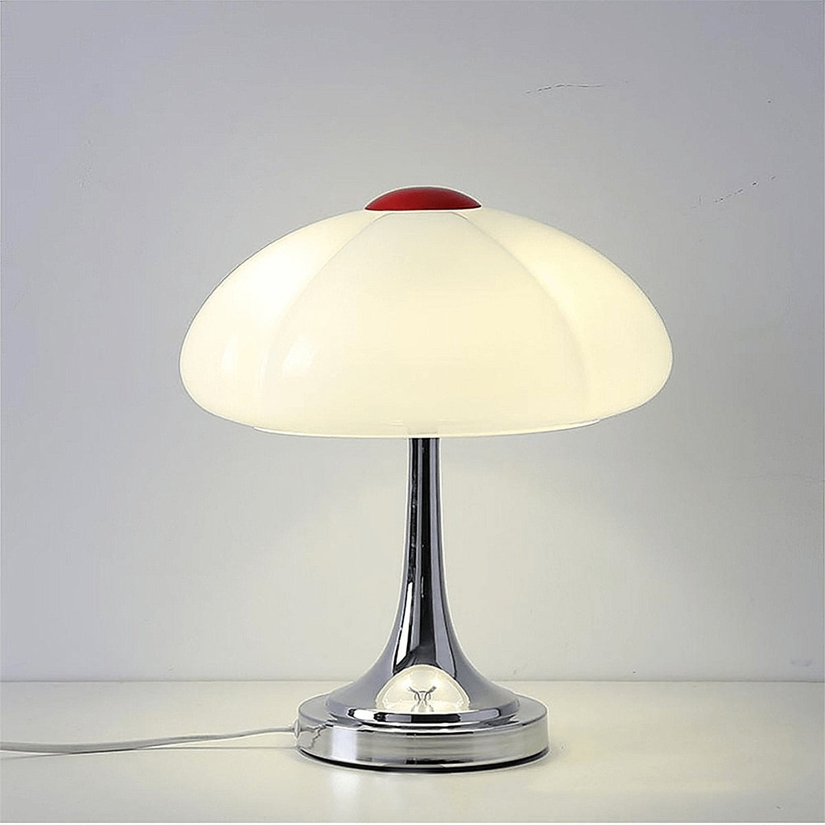 Close front view of this Clérachic lamp lit on a white surface, the creamy glass shade and red cap above a polished chrome base.