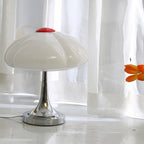 Unlit Clérachic table lamp in front of sheer white curtains, its white glass shade and red cap above the chrome stem.