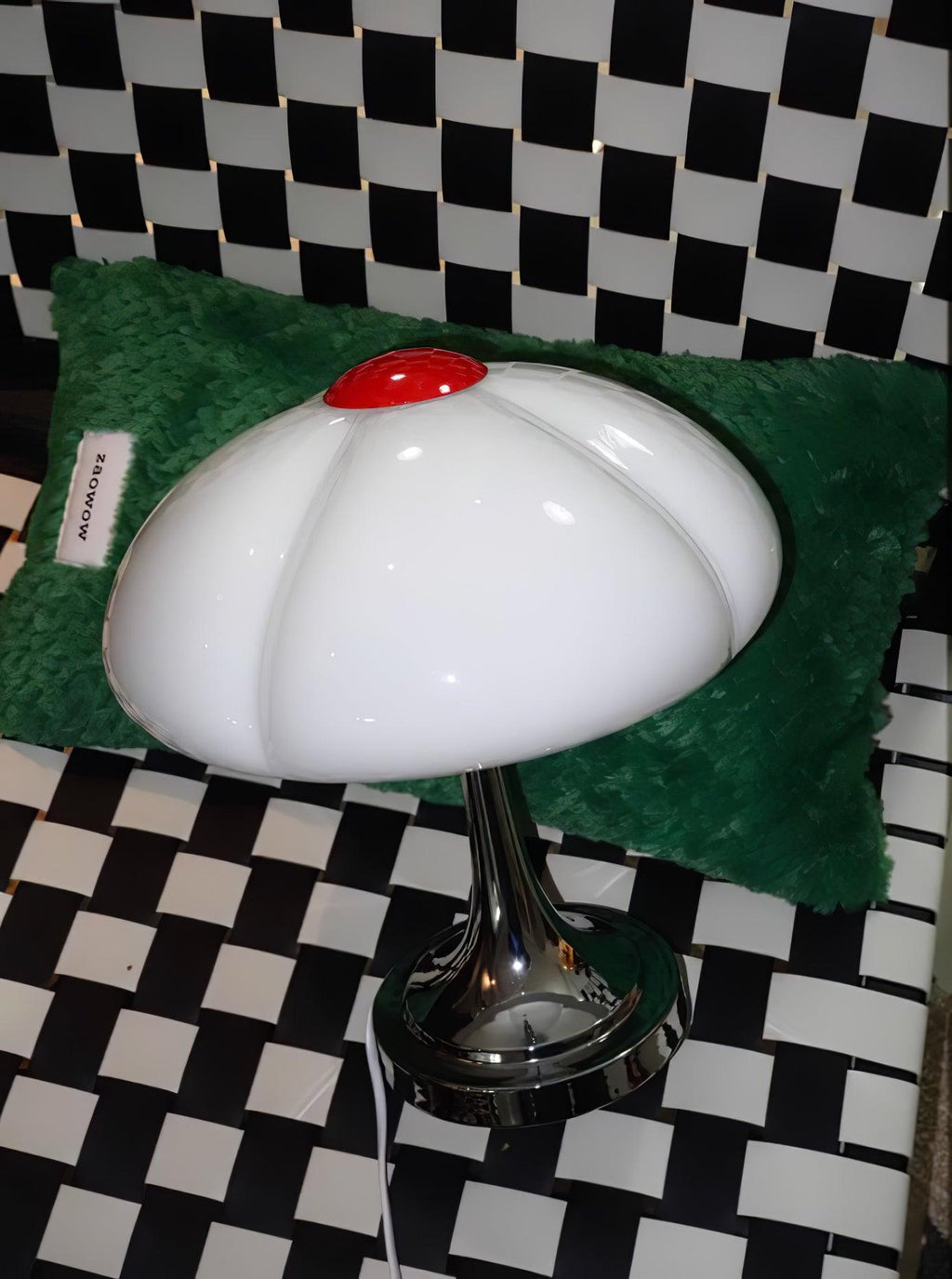 This Clérachic lamp on a black-and-white checked floor, in front of a green fluffy cushion, its glass shade unlit.