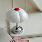 Clérachic table lamp resting on a cream cushioned bench, its white glass shade and chrome base beside a pink book.