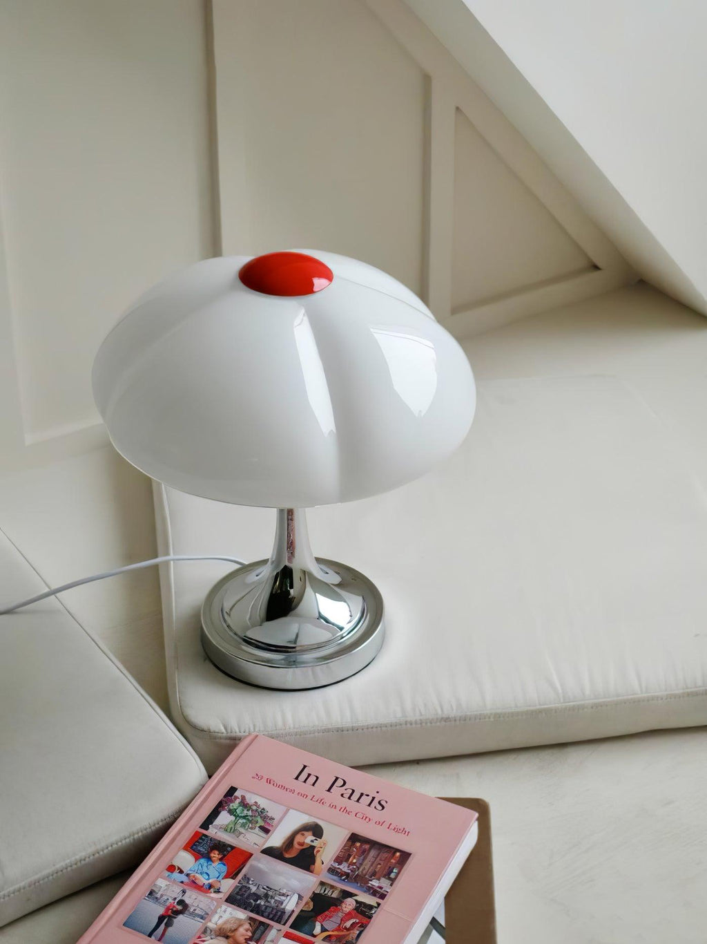 Pink book at the foot of this Clérachic lamp, which stands on a cream cushioned seat in a bright corner.