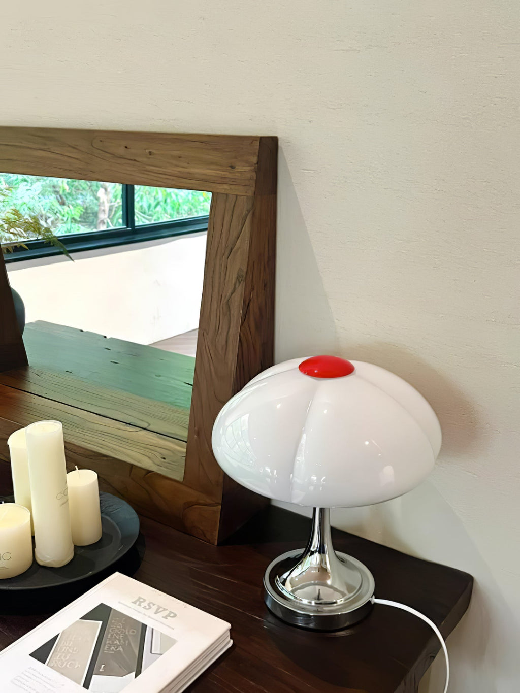 A white Clérachic table lamp in front of a wooden-framed mirror, with pillar candles and a book on a dark console.