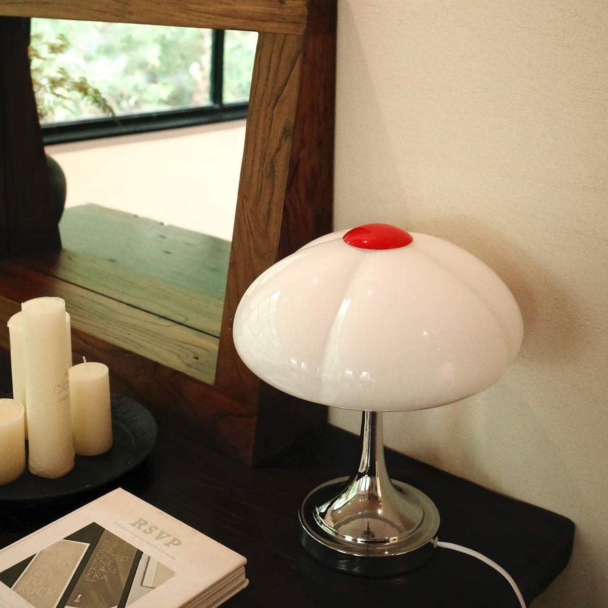 Clérachic table lamp with a white glass shade in front of a wooden mirror frame, above a dark console and an open book.