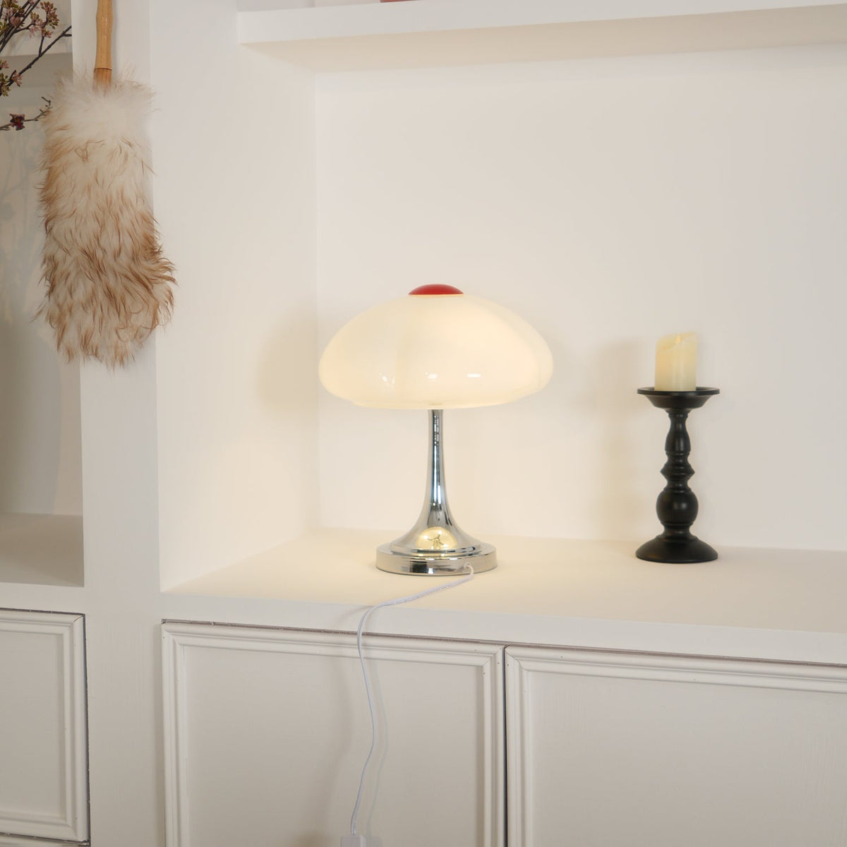 Warmly lit Clérachic lamp on a white shelf with a black candlestick, its cord hanging down over the cabinet doors.