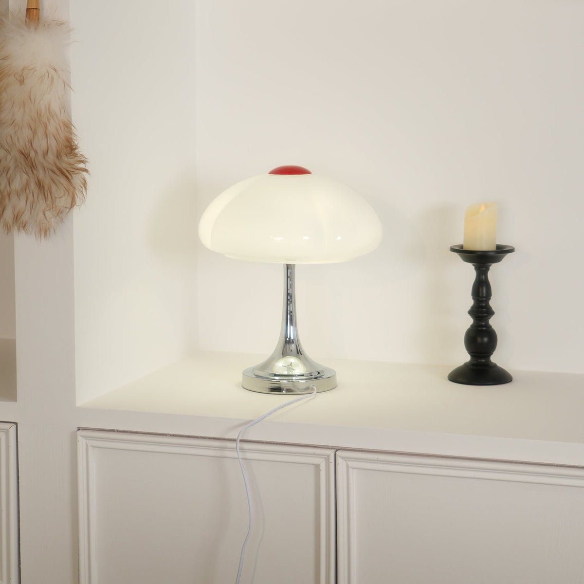 A softly lit Clérachic lamp on a white shelf beside a black candlestick, the shade glowing in a cream tone.
